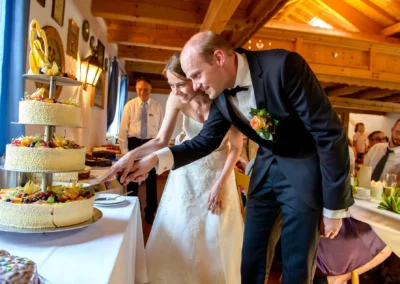 Paar schneidet Hochzeitstorte bei Hochzeitsfeier in Trachtenheim Irschenberg. Hochzeitsfotografie in action im Bild.