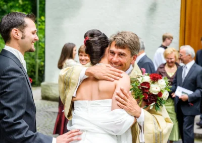 Priester umarmt Brautpaar vor Kirche bei Hochzeitsfeier in München. Gäste im Hintergrund, Blumenschmuck. Hochzeitsfotograf.