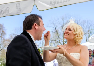 Braut fuettert Bräutigam mit Kuchen bei Hochzeit in Bad Toelz, lächelnd im Freien unter Sonnenschirmen.