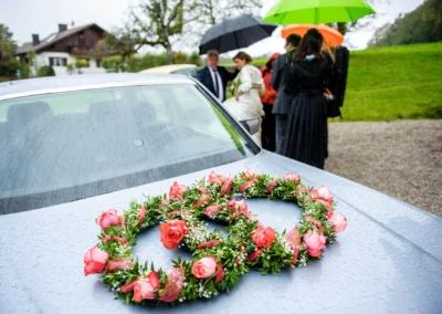 Auto mit Rosenkranz und Hochzeitsgesellschaft bei Regen. Hochzeitsfoto in Harkirchen, festgehalten von einem Hochzeitsfotografen.