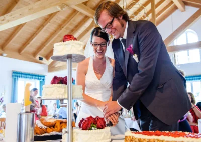 Brautpaar schneidet Hochzeitstorte bei Hochzeit nordlich von Dachau an. Fröhliche Gäste im Hintergrund. Hochzeitsreportage.