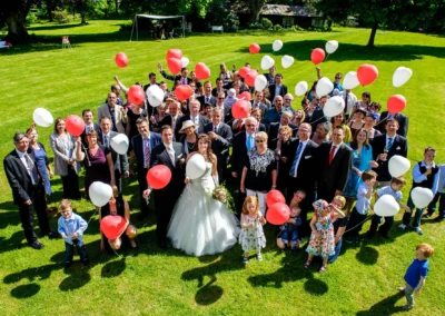 Hochzeitsfoto im Schloss Aufhausen: Gruppe von Gaesten mit roten und weissen Luftballons auf einer Wiese bei einer Hochzeit.