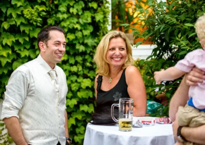 Paar lacht bei einer Hochzeit im Dachauer Hinterland, im Freien an einem Tisch mit Bierglas und Efeu im Hintergrund.