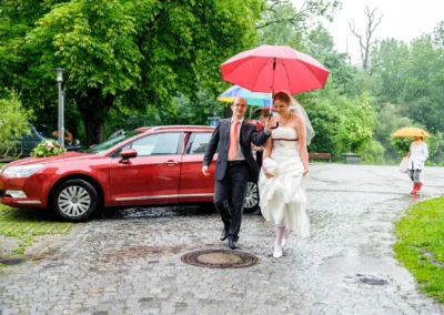 Brautpaar unter Regenschirm bei freier Trauung in Schloss Blutenburg. Rotes Auto im Hintergrund. Hochzeitsfotografie.