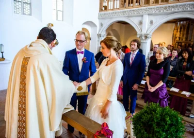 Paar und Priester bei kirchlicher Trauung in Landshut, Hochzeitsfotografie-Moment mit Gaesten im Hintergrund.