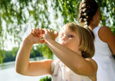Kind mit Weinglas bei Hochzeit am Langwieder See. Sonnenlicht strahlt durch Baumschatten. Hochzeitsfotografie.