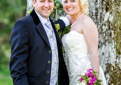 Brautpaar laechelt vor Baum im Kurpark Bad Toelz, Hochzeitsfoto in elegantem Stil, Oberbayern.