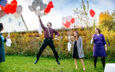 Drei Personen halten bunte Herzballons im Garten; einer springt freudig in die Luft.