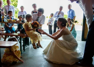 Braut ueberreicht Blumenmaedchen Strauß bei Hochzeit im Landkreis Miesbach, Gaeste im Hintergrund in festlicher Kleidung.