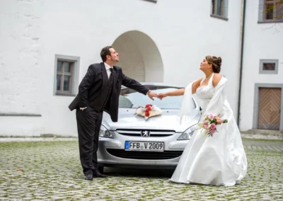 Brautpaar vor Auto mit Blumenstrauss, aufgenommen auf Hochzeit nahe Dillingen. Hochzeitsportrait mit romantischem Motiv.