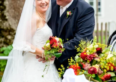 Brautpaar lacht vor klassischem Auto mit Blumenstrauss bei Hochzeit im Brauereigasthof Aying. Hochzeitsfotografie Aying.