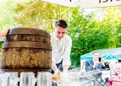 Mann zapft Holzfass bei Hochzeit in Schwabing, München. Gäste beobachten und fotografieren fröhlich. Hochzeitsfotografie.