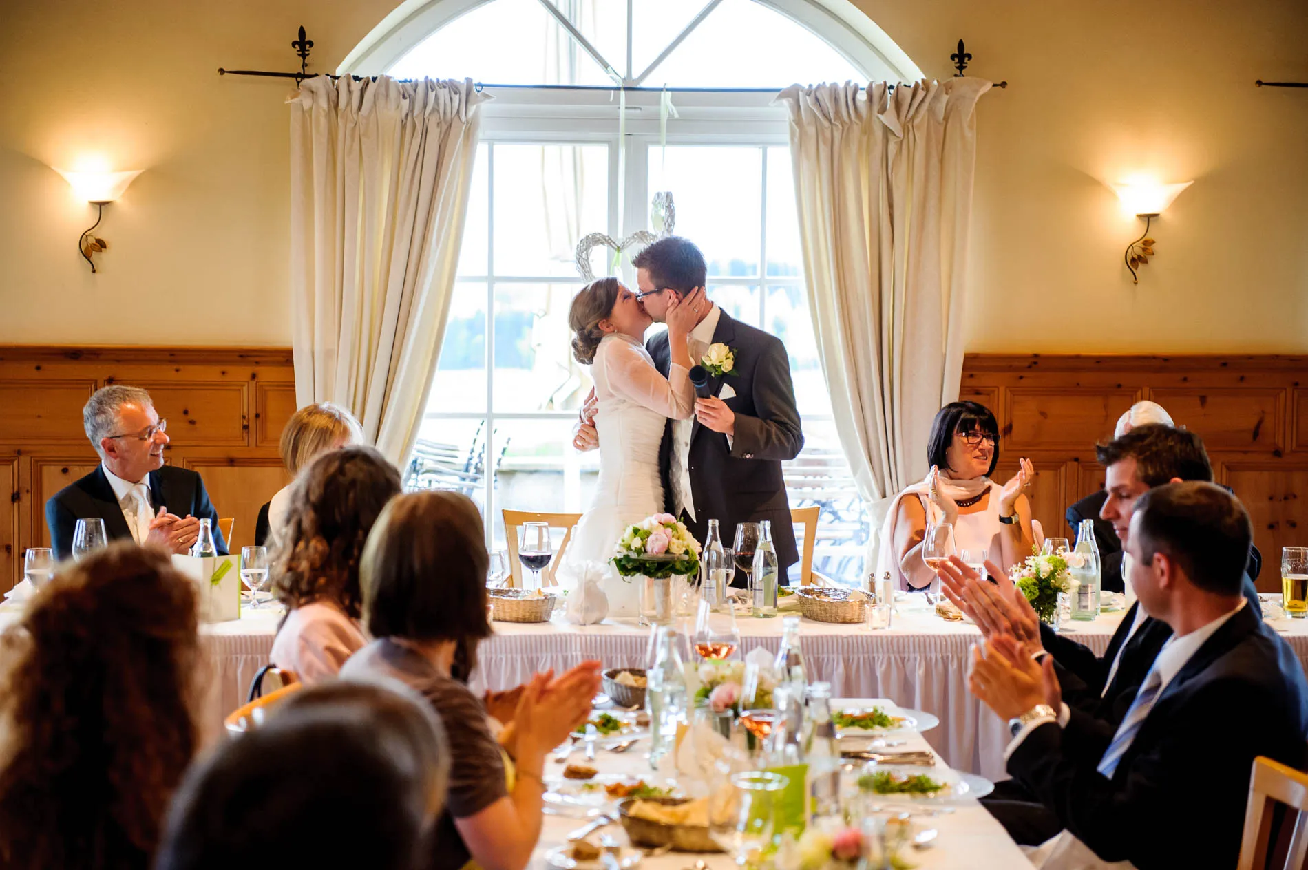 Brautpaar teilt Kuss bei Hochzeit im Aschbacher Hof, Gaeste applaudieren am Tisch. Hochzeitsfotografie in Bayern.