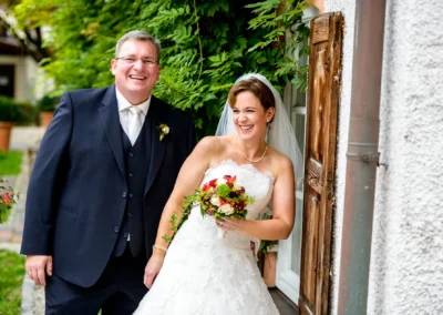 Bräutigam im Anzug und Braut im Kleid lachen bei der Hochzeitsreportage in Aying vor grünem Laub.