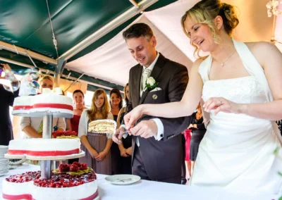 Brautpaar schneidet Hochzeitstorte an, umgeben von Gaesten in Schwabing, Muenchen. Hochzeitsfotografieaufnahme.