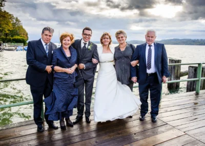 Brautpaar und Familie posieren bei der Hochzeitsreportage am Starnberger See auf Holzsteg mit Wolkenhimmel im Hintergrund.