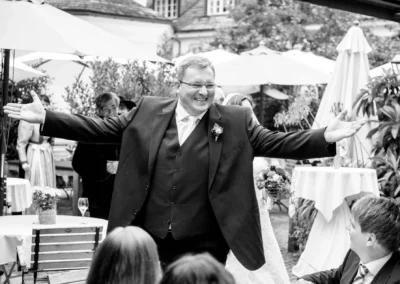 Mann in Anzug lacht herzlich bei Hochzeit im Freien in Aying. Hintergrund mit Gaesten und Tischen. Hochzeitsreportage.