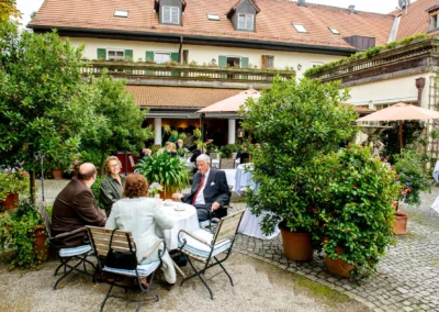 Gartenfest bei Hochzeit im Brauereigasthof Aying, Gaeste sitzen an Tisch im Freien surrounded von Pflanzen und Dekor.