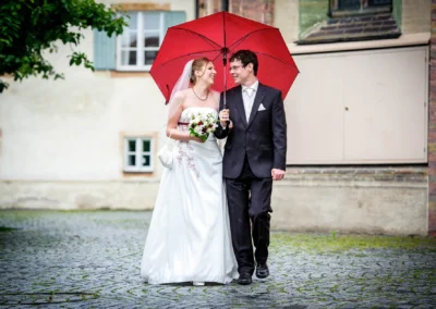 Brautpaar unter rotem Regenschirm bei Hochzeitsfotos im Schloss Blutenburg, lachend und verliebt.