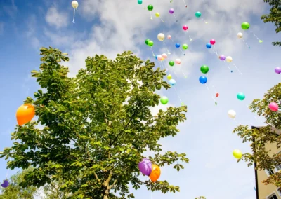 Bunte Luftballons steigen bei einer Hochzeit am Langwieder See in den Himmel, umrahmt von gruenen Baeumen.