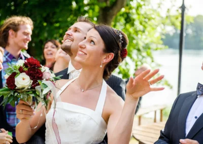 Brautpaar lacht bei Hochzeit am Langwieder See, umgeben von Gaesten und Baum im Hintergrund. Hochzeitsfotografie.