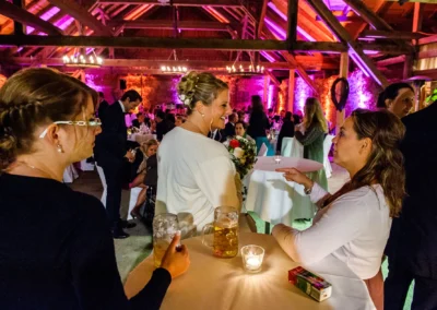 Gäste bei Hochzeit in Eventscheune Wallenburg sitzen an Tisch mit Getränken, warme Beleuchtung, festliche Stimmung.