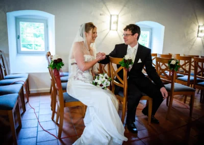 Brautpaar sitzt lächelnd nebeneinander in Hochzeitszeremonie in München, elegante Dekoration auf Stühlen.