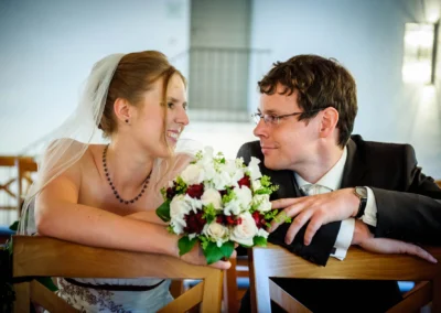 Brautpaar beim Hochzeitsportrait in einem Saal, lachend hinter Blumenstrauss. Hochzeitsfoto in München-Obermenzing.