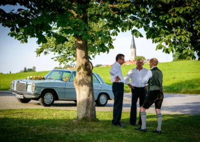 Drei Männer stehen unter einem Baum, Hochzeitsauto und Kirche im Hintergrund in irschenberg. Hochzeitsfotograf.