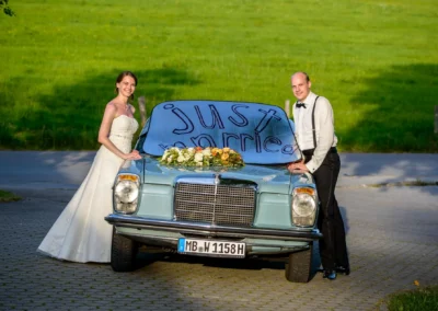 Brautpaar vor Oldtimer mit Just Married Schild, aufgenommen bei Hochzeit nahe Irschenberg. Hochzeitsfoto mit Blumen.