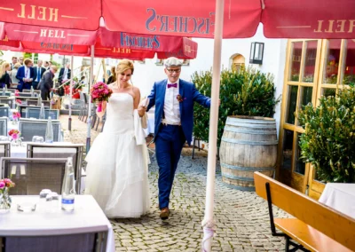 Brautpaar lacht und geht unter roten Sonnenschirmen bei Hochzeit in Landshut, umgeben von Blumen und Gaesten.