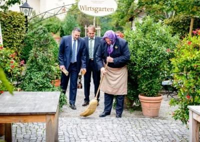 Hochzeitsfoto im brauereigasthof aying: Drei Maenner, einer fegt mit Besen, im gruenen Wirtshausgarten.