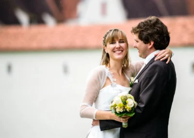 Brautpaar beim Hochzeitsfoto vor Schloss Grunau, Neuburg an der Donau, lachend und umarmt mit Blumenstrauss.