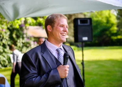 Mann im Anzug haelt Mikrofon bei Hochzeit im Freien, Schloss Aufhausen, Oberbayern. Hochzeitsfotografaufnahme.