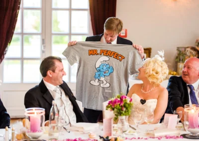 Hochzeit in Bad Toelz: Mann haelt lustiges T-Shirt hoch, Brautpaar lacht am Tisch bei der Feier. Hochzeitsfotografie.