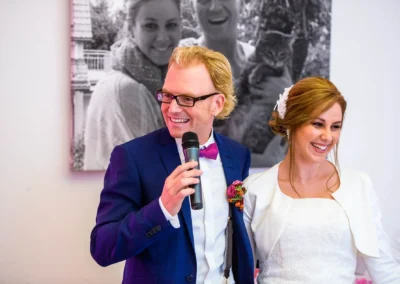 Bräutigam im blauen Anzug mit Mikrofon neben Braut in Weiss bei Hochzeit im Augustlhof Pfettrach, Hochzeitsfotografie.