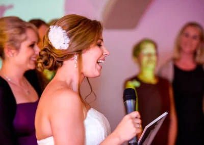 Braut bei einer Hochzeitsrede mit Mikrofon in Pfettrach bei Landshut. Hochzeitsfoto mit Freunden im Hintergrund.