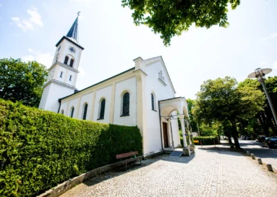 Eine weiße Kirche mit Turm, umgeben von Bäumen, daneben gepflasterter Weg und Grünfläche, sonniger Tag.