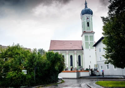 Kirche in Zorneding östlich von München mit Zwiebelturm, Regen, Person mit buntem Regenschirm, Bäume und ein Auto im Vordergrund.