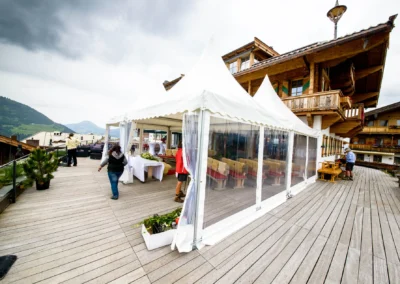 Ein Hochzeitspavillon auf einer Holzterrasse mit Berghütten, gedeckte Tische, Menschen bereiten Dekoration vor.
