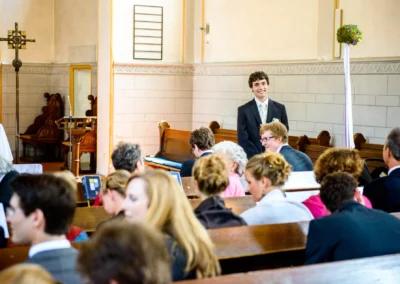 Ein lächelnder Bräutigam steht in einem Kirchenraum; Gäste sitzen in Bankreihen und warten erwartungsvoll.
