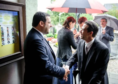 Zwei Männer in Anzügen schütteln sich lächelnd die Hände, während um sie herum Personen mit Regenschirmen stehen.