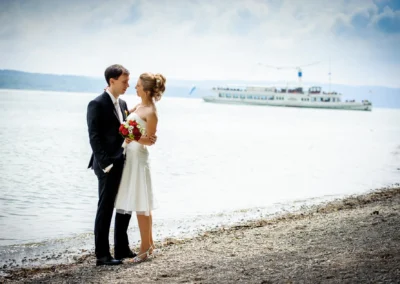 Brautpaar am Ufer des Ammersees, Hochzeitsfotografie im Landkreis Landsberg-Lech mit Schiff im Hintergrund.