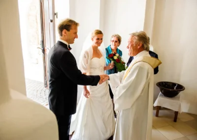 Bräutigam schüttelt Hand des Priesters, Braut mit Blumenstrauß lächelnd daneben. Hochzeitsfoto Kirchheim bei München.