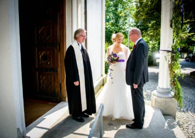 Braut in weißem Kleid mit lila Blumenstrauß, lächelt neben älterem Paar und Geistlichem vor geschmücktem Kircheneingang.