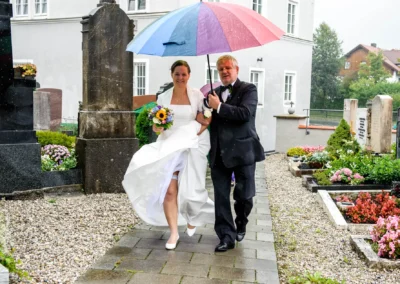 Die Braut in Weiß und ein Mann gehen unter einem bunten Regenschirm, dabei hält sie einen Blumenstrauß.