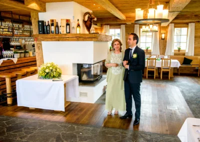 Ein Mann im Anzug mit Ansteckblume und eine Frau in grünem Kleid stehen in einem rustikalen Raum neben einem Tisch mit Blumen.