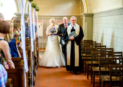 Eine Braut im weißen Kleid mit Blumenstrauß betritt die Kirche, begleitet von einem Mann im Anzug und einem Geistlichen.