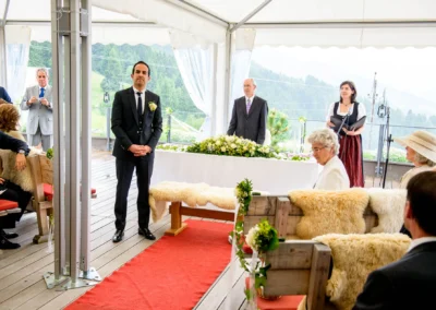 Ein Bräutigam im Anzug steht unter einem Zelt, umgeben von Gästen auf Holzbänken mit Fellauflagen, und Blümendekoration.