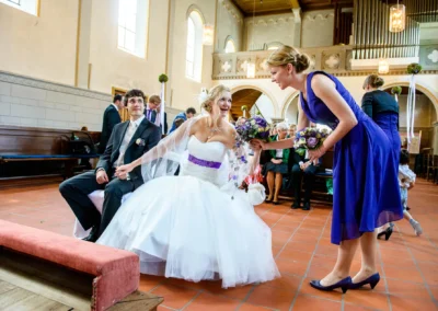 Das Brautpaar sitzt in der Kirche in Starnberg. Die Braut im weißen Kleid mit lila Band lächelt. Sie hält Händchen mit dem Bräutigam.